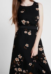 Nainen ilman hihnoja mustassa mekossa, jossa on beige- ja ruskeakuvioisia kukkia, joustava vyötärö, pitkä ruskea hius ja käsi hameen päällä.