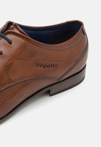 Chaussure habillée en cuir marron avec un design perforé, lacets noirs et intérieur bleu contrastant. Semelle robuste avec un léger talon.