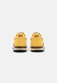 Des chaussures de sport jaunes avec des accents marron et une surface texturée, dotées d'un col rembourré et du logo ASICS sur le talon.
