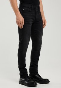 Férfi fekete slim-fit farmernadrágban és fekete bőr cipőben, áll egy egyszerű világos háttér előtt.