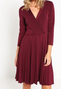 Burgunderfärgad klänning med en plisserad kjol, omlottskärning och trekvartsärmar. Tillverkad av ett mjukt tyg med en slät textur.