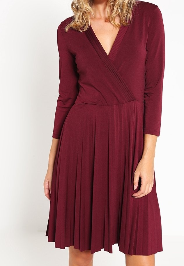 Burgunderfärgad klänning med en plisserad kjol, omlottskärning och trekvartsärmar. Tillverkad av ett mjukt tyg med en slät textur.