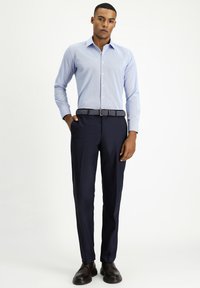 Camicia a righe blu chiaro con colletto a bottoni, abbinata a pantaloni navy. Cintura intrecciata aggiunge un contrasto. Il modello sta in piedi con le mani in tasca.