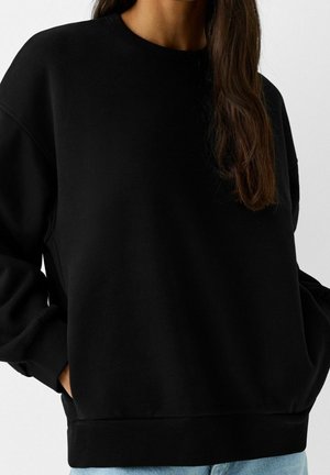 Sweatshirt noir oversize avec des manches longues, un col et des poignets côtelés, et un tissu texturé. Comprend des poches latérales et une coupe décontractée.