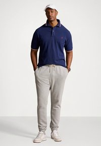 Μπλε σκούρο πόλο μπλουζάκι με κόκκινες λεπτομέρειες, συνδυασμένο με ανοιχτό γκρι φόρμες και λευκά αθλητικά παπούτσια, φορεμένο από έναν άνδρα που στέκεται.