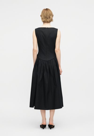 Osoba o krótkich blond włosach ubrana w czarną, bezrękawnikową sukienkę midi z dopasowaną górą i rozkloszowanym dołem, widziana od tyłu.