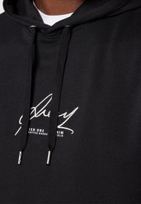 Sort hættetrøje lavet af blødt stof, med stort hvidt logo og tekst på fronten. Snore med sølvfarvede metalender fuldender designet.