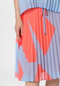 Gevouwen rok in blauw en koraal, met geometrische patronen. Licht, vloeiend materiaal met een trekkoorddetail en een gestructureerde uitstraling.