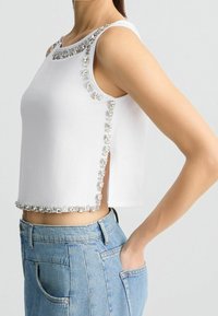 Top bianco corto con applicazioni di cristalli lungo lo scollo e i lati. Abbinato a jeans a vita alta di colore blu chiaro.