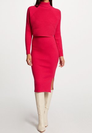 Kobieta ubrana w dopasowaną, jasnoróżową prążkowaną sukienkę z długimi rękawami i rozcięciem z boku, w zestawieniu z białymi kozakami do kolan.