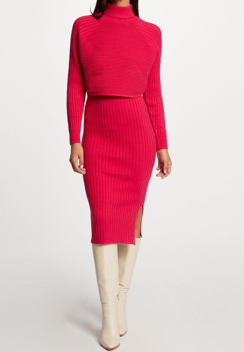 Femme portant une robe en maille côtelée rose vif ajustée, à manches longues avec une fente latérale, assortie à des bottes blanches montant jusqu'aux genoux.