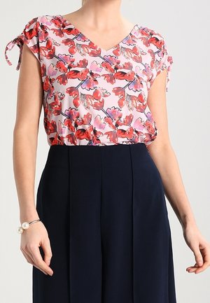 Kobieta ubrana w różową kwiecistą bluzkę z dekoltem w serek, z krótkimi rękawami zawiązanymi na węzeł, i wysokim stanem ciemnoniebieskich spodni, na jednolitym tle.