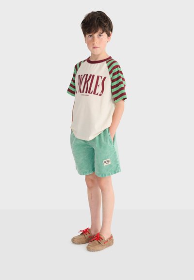 Garçon debout les mains dans les poches, portant un t-shirt à manches courtes rayé rouge et vert, un short vert, et des chaussures marron avec des lacets rouges.