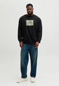Sort sweatshirt med en rektangulær logo-patch, kombineret med løstsiddende blå jeans og hvide sneakers, stående mod en neutral baggrund.