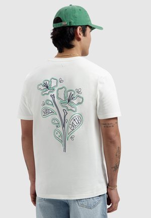 Man draagt een wit T-shirt met een groen omrande bloemdesign en de tekst 'live' en 'faith' op de achterkant, een groene pet, lichtblauwe spijkerbroek, kijkt naar achteren.