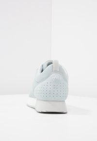 Chaussure de sport bleu clair avec une tige en mesh et daim, un talon perforé, et un logo blanc sur la languette arrière. Semelle en caoutchouc avec motif antidérapant.