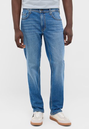 Mann trägt mittelblaue Jeans mit geradem Bein und weiße Sneakers vor einem schlichten hellen Hintergrund.
