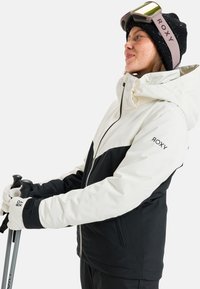 Kurtka narciarska w białym i czarnym kolorze, z zamek błyskawicznym z przodu, elastycznymi mankietami i logo Roxy. Rękawice trzymają kije narciarskie na jednolitym tle.