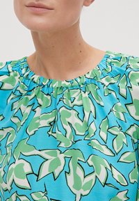 Haut bleu clair avec un col froncé, présentant un motif de feuilles vertes et blanches surligné de noir. Le tissu a une texture douce et lisse.