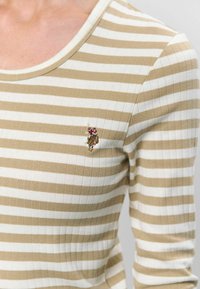 Kremskrāsas un baltas svītrainas ribotas garām piedurknēm blūze ar apļu kakla izgriezumu, kam pie krūts ir neliels izšūts modes figūras akcents.