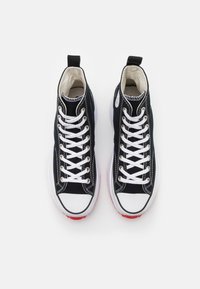 Paire de baskets montantes noires en toile avec lacets blancs, bouts en caoutchouc et semelles rouges vues de dessus sur un fond blanc.