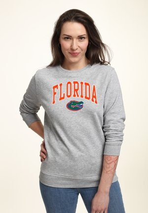 Frau mit langen dunklen Haaren, die einen grauen Pullover mit Florida-Schriftzug und Alligator-Logo trägt, steht mit der Hand in der Hüfte und einem Tattoo am Unterarm.