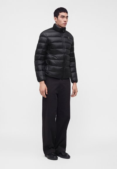 Homme debout portant une doudoune noire, un pantalon noir à jambes larges et des chaussures noires sur un fond uni gris clair.