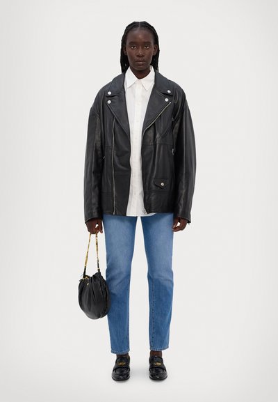 Mustmust nahkainvestus hõbedaste detailidega, valge nööbiga särk, helesinised sirged teksad ja mustad loafers'id, hoides musta kotti.