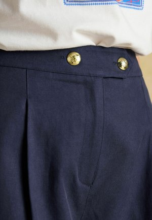 Marineblaue Hose mit Falten und Seitentaschen, mit goldenen Knöpfen am Bund. Der Stoff wirkt glatt mit einer strukturierten Passform.
