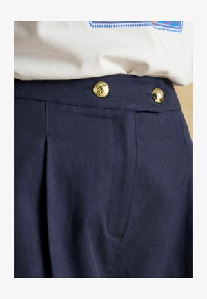 Marineblaue Hose mit Falten und Seitentaschen, mit goldenen Knöpfen am Bund. Der Stoff wirkt glatt mit einer strukturierten Passform.