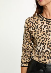 Un suéter de leopardo en beige con manchas negras, textura acanalada y cuello negro. Las mangas son largas y ajustadas, se lleva con pantalones negros.