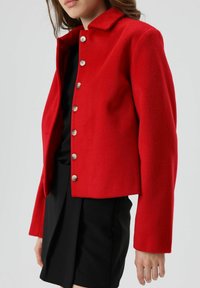 Rode jas van een wolblend met een kraag en zilveren knopen, met een kort design en een getailleerd silhouet. Draagt over een zwarte rok.