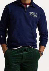 Marinblå zip-up sweatshirt med framficka och broderad text "POLO RALPH LAUREN". Har ribbade ärmslut och krage. Matchas med gröna kordbyxor.