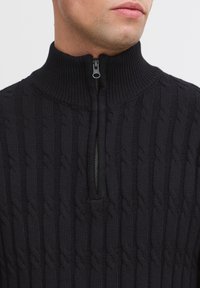 Zwarte gebreide pullover met een hoge kraag met ritssluiting. Heeft een geribbelde textuur en kabelbreipatroon, wat zorgt voor een comfortabele pasvorm en een casual uitstraling.
