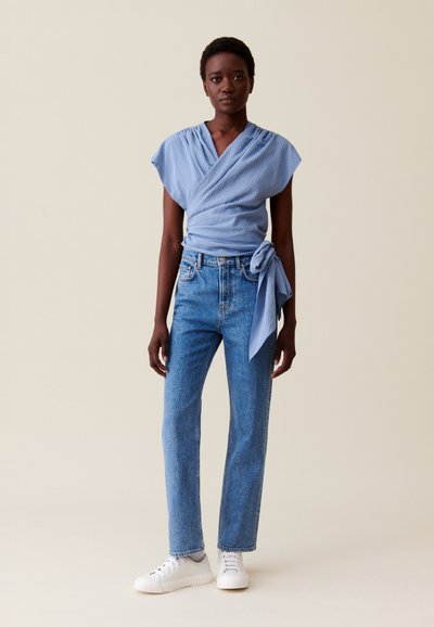 Top drapé bleu clair à manches courtes, associé à un jean bleu taille haute à jambe droite et des baskets blanches avec des accents gris.