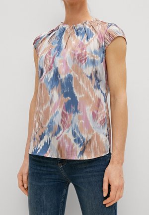 Kvinde iført en ærmeløs, plisseret bluse med abstrakt mønster i pink, blå, beige og hvid, kombineret med mørkeblå jeans.