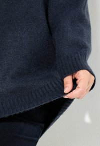 Dłoń trzymająca brzeg granatowego dzianinowego swetra, ukazująca teksturę oraz ribbowe wykończenie rękawa i dolnej krawędzi.