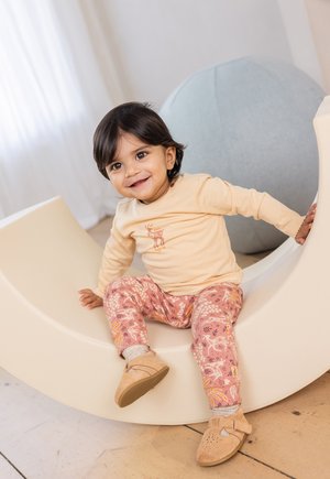 Hellbeige Langarmshirt mit einem Reh-Design, rosa gemusterte Hose mit blumigen Motiven und sandfarbene Schuhe an einem Kind, das auf einem weißen, geschwungenen Stuhl sitzt.