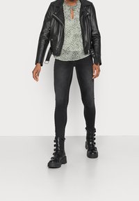 Giacca da moto in pelle nera sopra una blusa verde chiaro con collo a cravatta, abbinata a jeans skinny neri e stivali neri chunky con dettagli a fibbia.