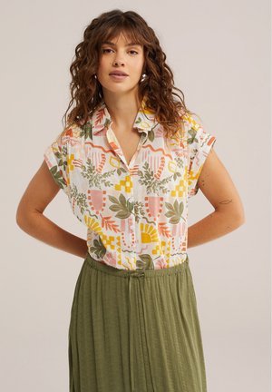 Žena oblečená v krátkosleevej košeli s farebnými abstraktnými kvetinovými vzormi a olivovo zelenou sukňou, stojaca s rukami za chrbtom.