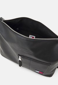 Bolsito de cuero negro con una cremallera brillante, bolsillo interior y el logo de Tommy Hilfiger. Presenta un diseño plano y una textura suave.