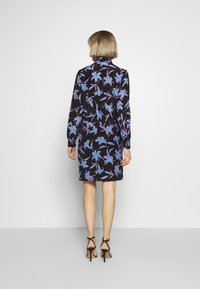 Modella che indossa un abito floreale nero con motivi blu e rosa, rivolta di spalle, in piedi su uno sfondo neutro, con sandali con tacco alto.
