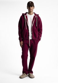 Sweat à capuche bordeaux avec des cordons blancs, associé à un pantalon de survêtement bordeaux assorti. T-shirt blanc en dessous, porté avec une casquette marron et des chaussures beiges.