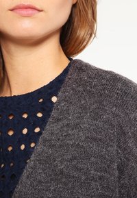 Grå stickad tröja bredvid en marinblå texturerad topp med utskärningsmönster, som visar en närbild av materialet och design detaljer.