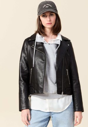 Veste en cuir noir de motard avec des zips argentés, superposée à un sweat à capuche gris et une chemise blanche, assortie d'une casquette grise.