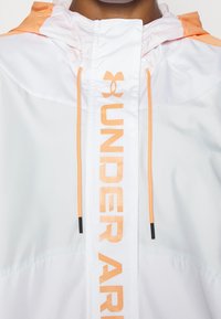 Veste à capuche blanche avec des accents orange et des cordons de serrage, arborant un logo "UNDER ARMOUR" audacieux sur le devant. Matériau léger et lisse.