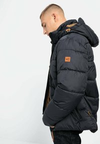 Schwarze Daunenjacke mit Kapuze, ausgestattet mit einem braunen Innenfutter und sandfarbenen Akzenten. Auffällig durch ihre gesteppten Texturen und Reißverschlusstaschen.