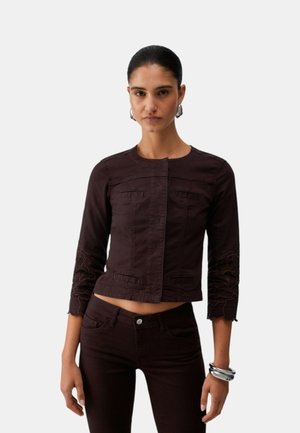 Donna con capelli scuri raccolti in uno chignon ordinato, che indossa una giacca aderente marrone scuro con maniche tre quarti e pantaloni abbinati, accessoriata con orecchini a cerchio d’argento e braccialetti.