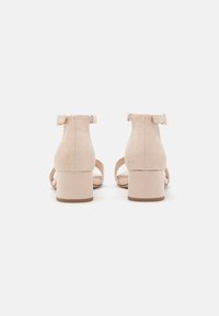 Beige mokkasandaler med hæl, åben bag, og ankelrem. Teksturen er blød, med en glat finish og et minimalistisk design.