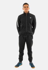 Zwarte fleece trainingspak met een ritsjack en broek, met witte strepen op de mouwen en een logo op de borst. Draagt met donkere sneakers en oranje accenten.